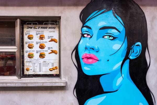 Graffiti image on a wall of a blue woman with pink lips next to a window with the burger menu of 3pm cafe.